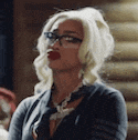 a woman with blonde hair and glasses is standing in front of a wooden wall .