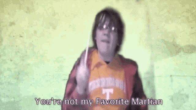 a man in an orange shirt with the letter t on it holds a knife and says you 're not my favorite martian