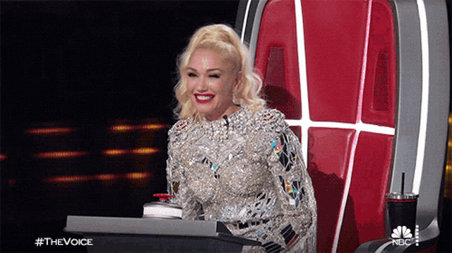 a woman in a silver dress is sitting in a chair with a nbc logo in the background