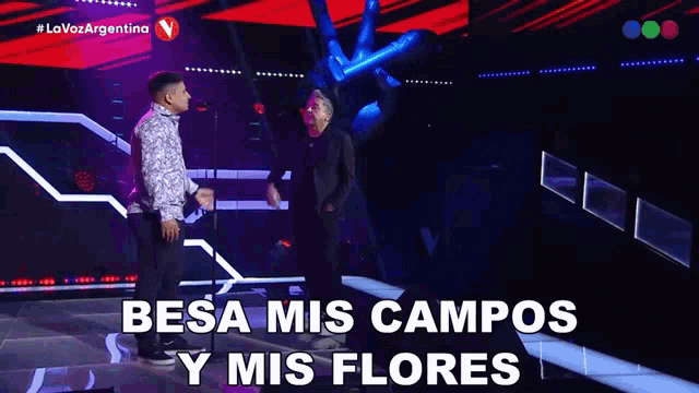 two men on a stage with the words " besa mis campos y mis flores " above them