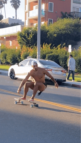 a shirtless man is riding a skateboard down a street with a white car behind him
