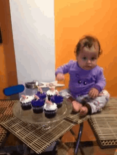 a baby in a purple shirt sits on a table with cupcakes on it