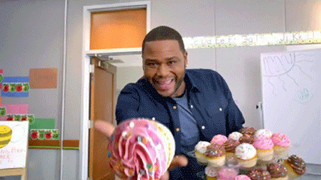 a man is holding a donut in front of a display of cupcakes in a classroom .