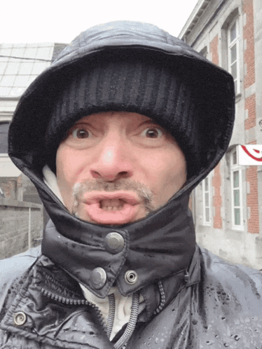 a man wearing a hooded jacket and a beanie makes a funny face in front of a post office