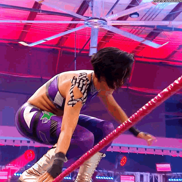 a wrestler in a purple outfit is jumping over a red rope with a fan in the background