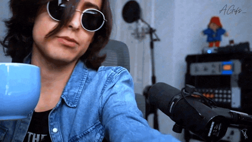 a man wearing sunglasses and a denim jacket holds a blue mug