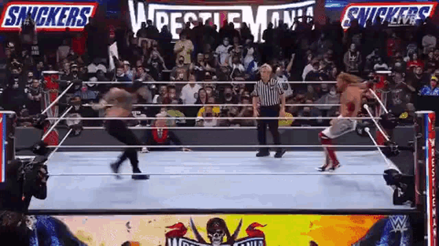 a wrestler is jumping over a referee in a wrestling ring during a match sponsored by snickers .