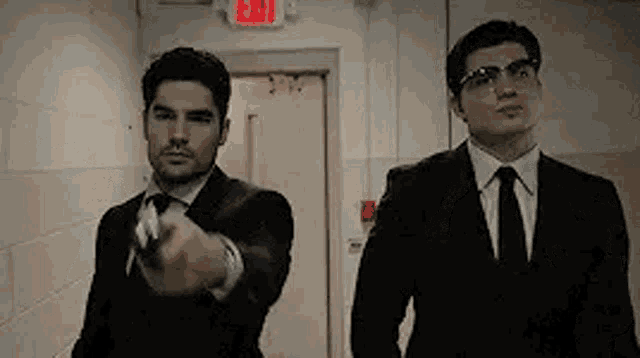 two men in suits and ties are standing next to each other in a hallway . one of the men is pointing a gun .