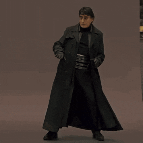 a man in a long black coat is standing on a brown background