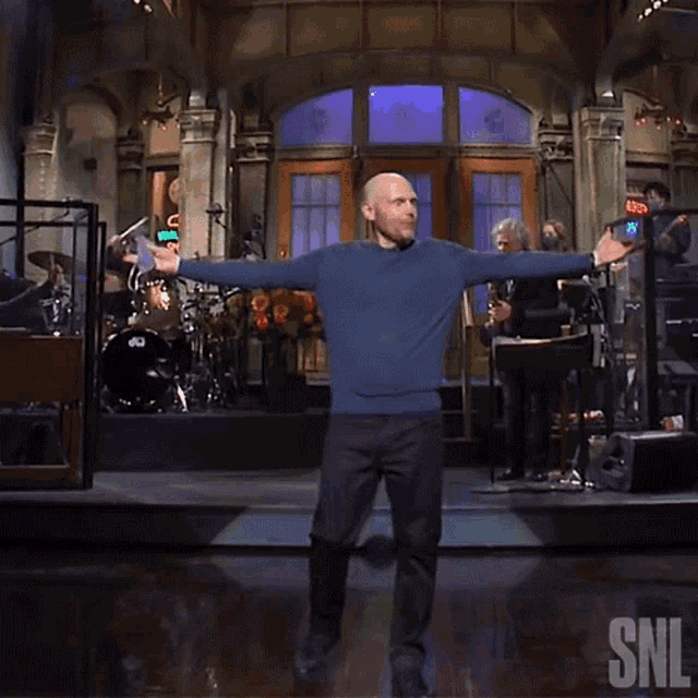 a man in a blue sweater stands on a stage with his arms outstretched in front of a snl logo