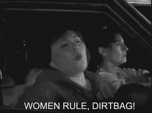 a woman is smoking a cigarette while driving a car with the words `` women rule dirtbag '' written on the screen .