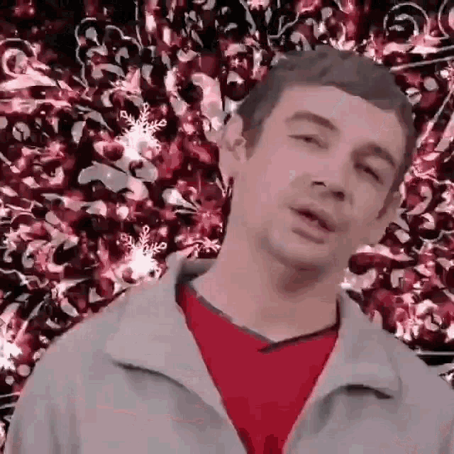 a man in a red shirt and gray jacket is standing in front of a christmas tree and making a funny face .