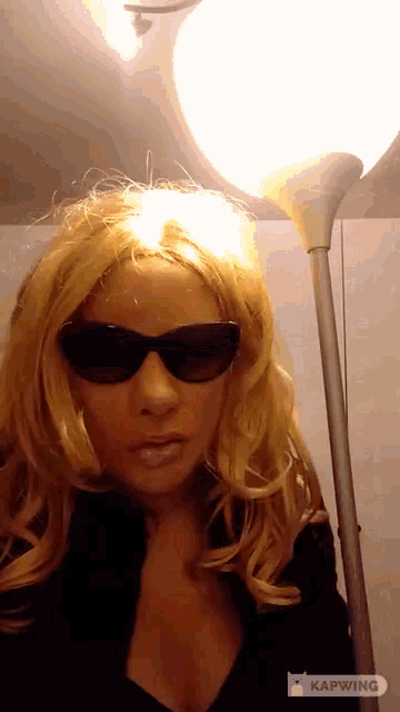 a woman wearing sunglasses and a wig is standing in front of a lamp with the caption kapwing
