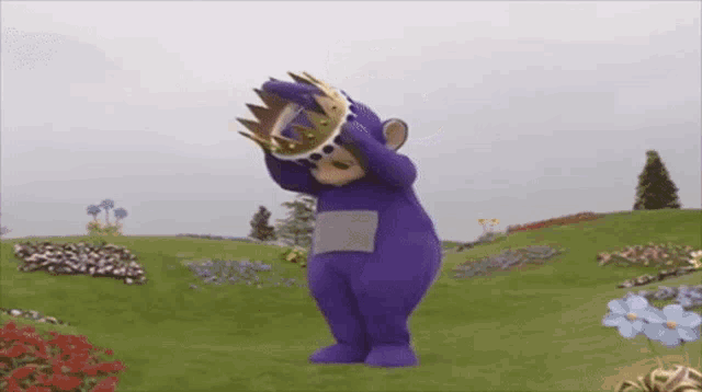 a purple teddy bear with a crown on his head is standing in a field of flowers