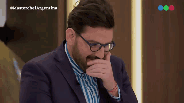 a man in a suit and glasses is covering his mouth while watching a masterchef argentina show