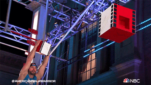 a man in a blue tank top is hanging upside down from a nbc sign