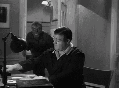 a man is sitting at a desk in front of a lamp while a werewolf is standing in the background .