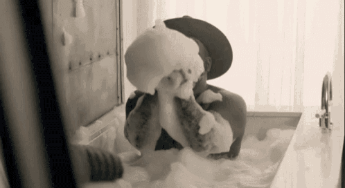 a man in a hat is taking a bath in a tub filled with foam .