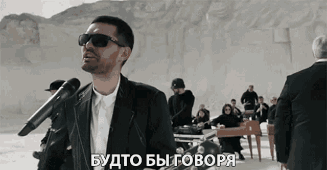 a man wearing sunglasses is singing into a microphone in front of a group of people in suits