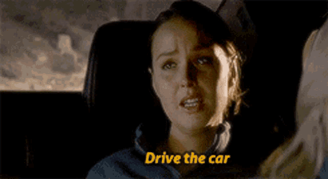 a woman is sitting in the driver 's seat of a car with the words `` drive the car '' written above her .