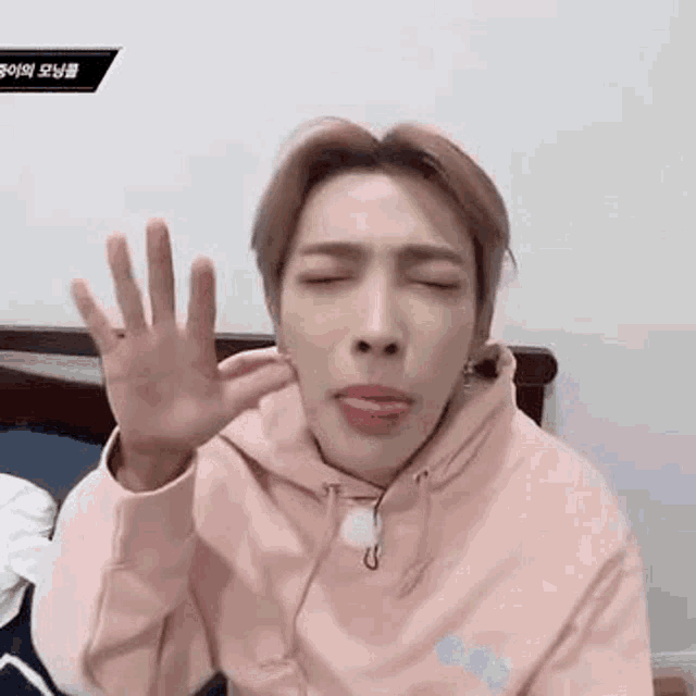 a man in a pink hoodie is sitting on a bed with his eyes closed and making a funny face .