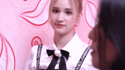 a woman wearing a white shirt with a black bow and dolce gabbana straps looks at the camera .