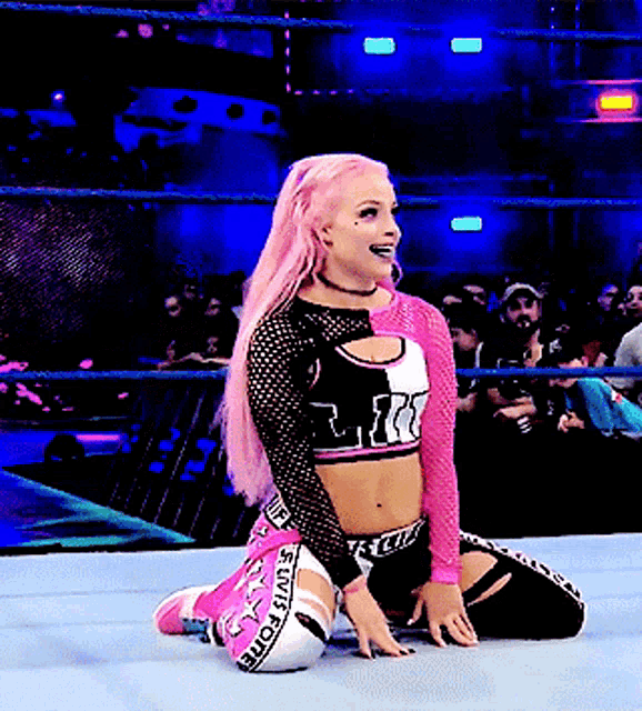 a woman with pink hair is kneeling on the floor in a wrestling ring .