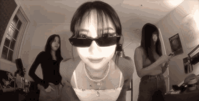 a woman wearing sunglasses and a pearl necklace stands in a room with two other women