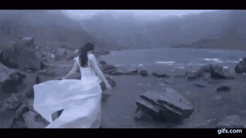 a woman in a white dress is walking on a rocky shoreline near a lake .