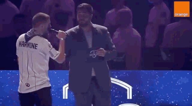 a man in a suit stands next to a man in a white shirt that says karmine on the back