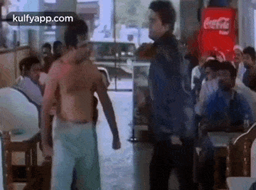 a man without a shirt is standing next to another man in a restaurant with a coca cola machine in the background .