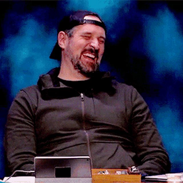 a man with a beard wearing a baseball cap is sitting in front of a tablet and smiling