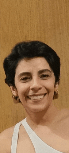 a woman wearing a white one shoulder top and hoop earrings is smiling in front of a wooden wall .