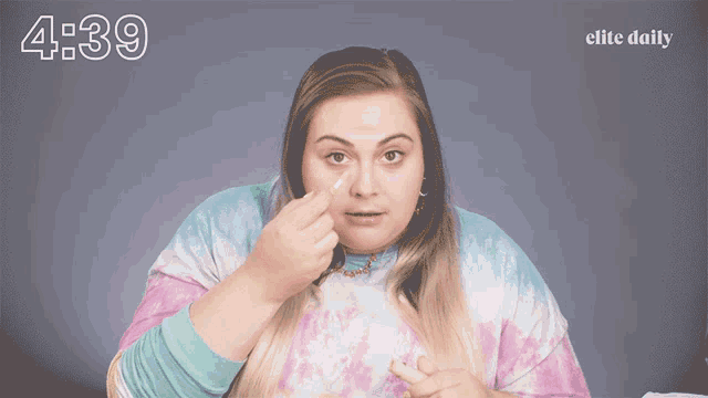 a woman wearing a tie dye shirt is applying a concealer to her face