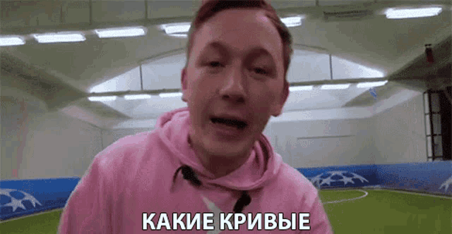 a man in a pink hoodie is standing in front of a soccer field and talking in a foreign language .
