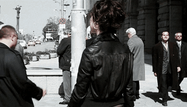 a woman in a black leather jacket stands in front of a pole with a no parking sign on it