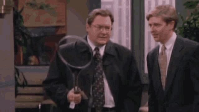 two men in suits and ties are standing next to each other . one man is holding a tennis racket .