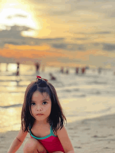 a little girl in a pink swimsuit sits on the beach at sunset