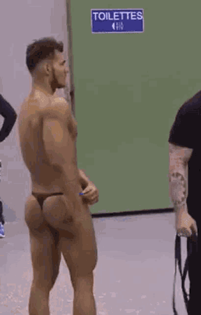 a shirtless man in a thong is standing in front of a blue sign that says toilettes .
