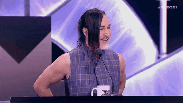 a man with long hair and a mustache stands in front of a microphone with # x2021 written on the screen behind him