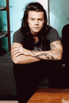 a man with long hair is sitting with his arms crossed and has a tattoo on his arm