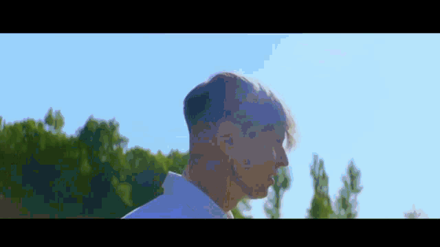 a man in a white shirt is standing in front of a blue sky and trees .