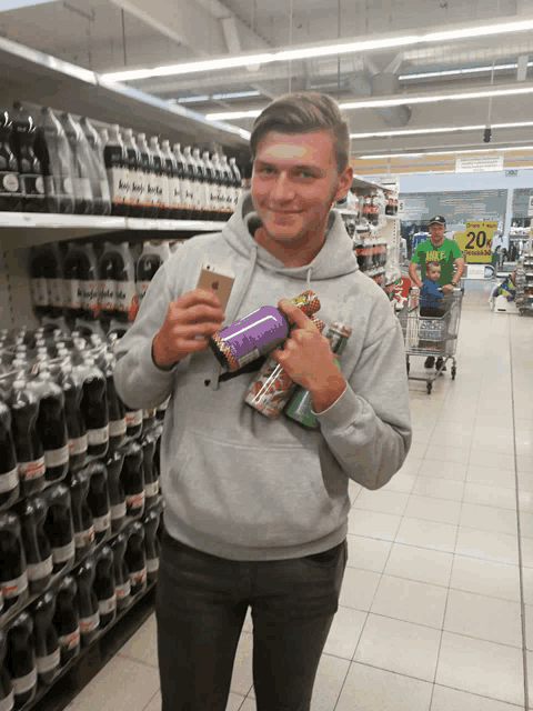 a man in a grey sweatshirt is holding a purple bottle and a can in front of a sign that says 20