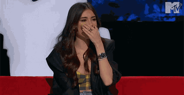 a woman is covering her mouth with her hand while sitting on a red couch in front of a mtv logo .