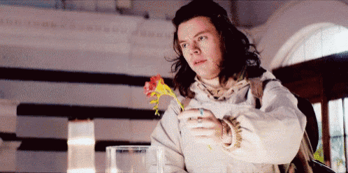 a man with long hair is holding a flower in his hand and looking at it .