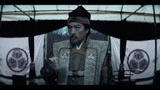 a man in a samurai costume is sitting in a tent with a flag in the background .