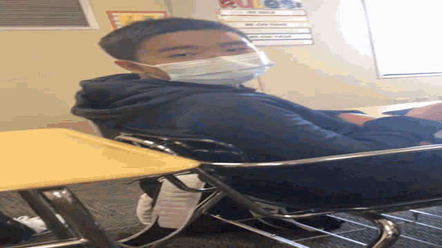 a boy wearing a mask sits at a desk in front of a bulletin board with rules on it