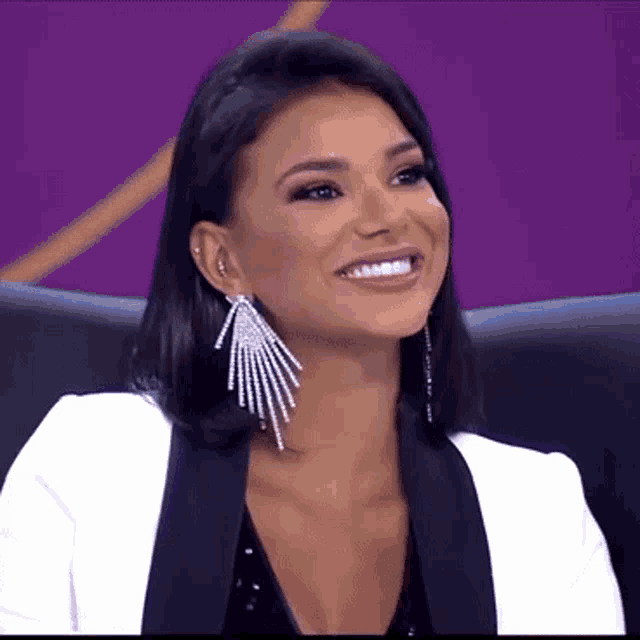 a woman wearing a white jacket and earrings is smiling and sitting in a chair .