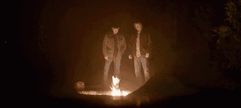 two men are standing around a campfire in the dark .