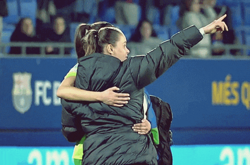 two women hugging on a soccer field with a fc barcelona banner in the background
